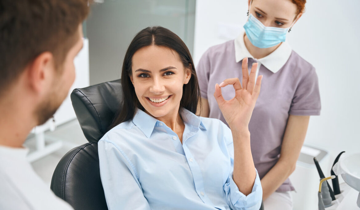 Smiling beautiful woman showing ok gesture sign sitting in orthodontic chair after painless and quality dental treatment, qualified dentist and nurse standing nearby