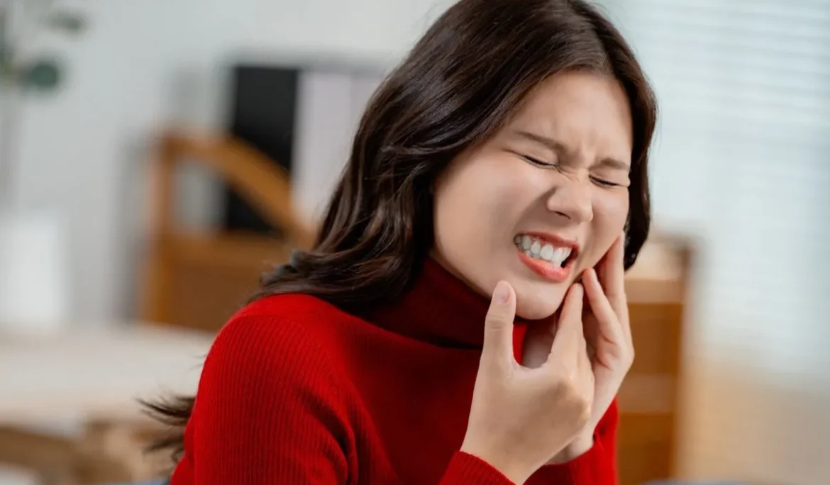 Asian woman having toothache, touching outside mouth with fingers, feeling painful, sitting on sofa at home